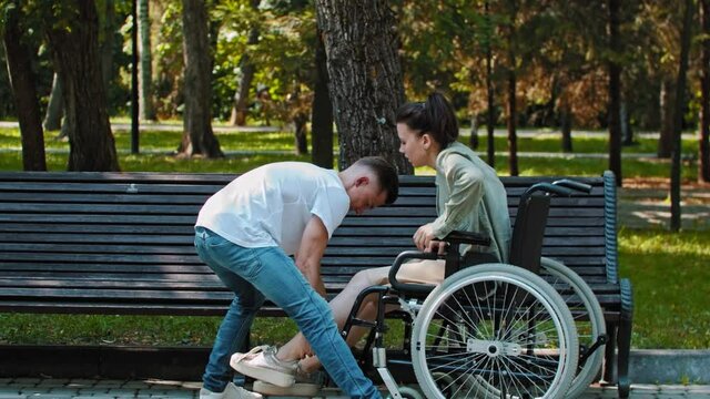 A couple walking in park - a guy transfers woman from a wheelchair to bench
