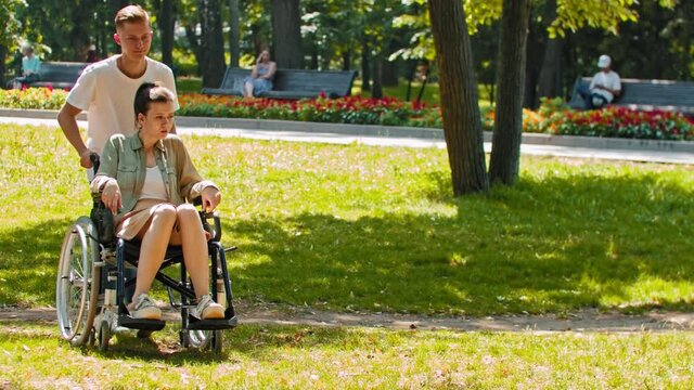 Friends in park - a guy rides disabled woman in a wheelchair then stops and sits next to her on the ground