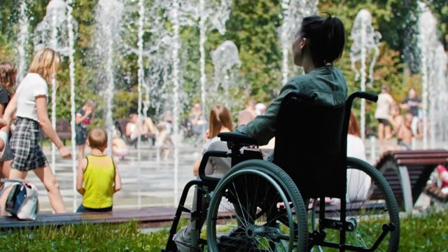 Sad woman in a wheelchair sitting in front of a fountain and looking at happy people