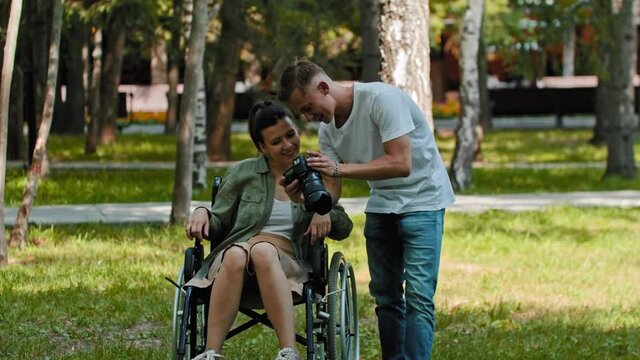 Woman in a wheelchair and her friend spending time at the summer park and the man showing her pictures from a camera