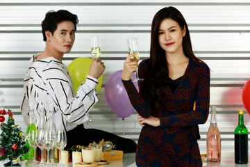Studio shot of Asian male pouring white wine into tall glass for female friend in fashionable wears standing together in celebration party in front round and star shape balloons on yellow background
