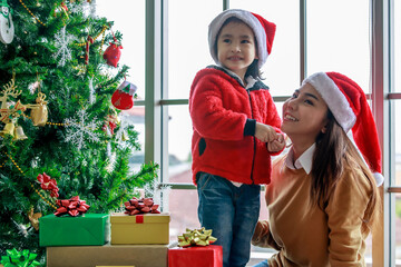 Happy Asian family mom and daughter in red and white Santa Claus hat and sweater smiling playing together near Christmas pine tree with glitter decorating items and present gift boxes in xmas evening