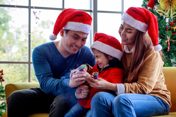 Happy Asian family father mother sitting on sofa helping afraid daughter wears sweater with red and white Santa Claus hat pulling exploding party popper confetti together celebrating Christmas eve