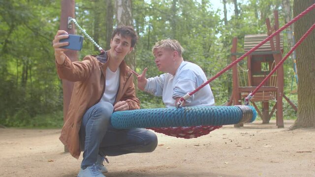 A man takes a selfie with a disabled person in a city park. He is holding a phone, they are posing, they are using the front camera. Image transmission at a distance.