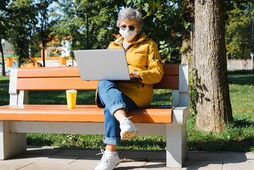 Alternative office, outdoor freelance. Senior woman in glasses and protective face mask browses Internet on laptop, looks at the screen, works while sitting on park bench. Older people and technology
