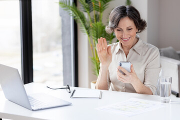 Portrait of elderly businesswoman sitting at an office or home desk use a mobile phone to communicate via the Internet. Technology, video call, online conference concept