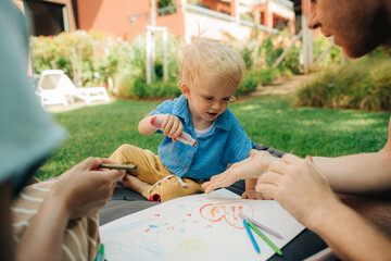 Funny little child painting with parents outdoors. Cute toddler girl resting with family on mattress at backyard. Parenthood and creativity concept