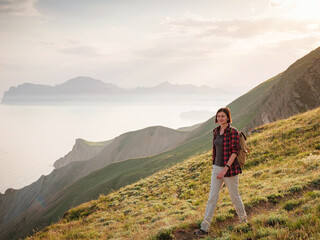 Fototapeta na wymiar A young Asian woman with a backpack hiking in the summer climbing the mountain at sunset . Mountain and coastal travel, freedom and an active lifestyle