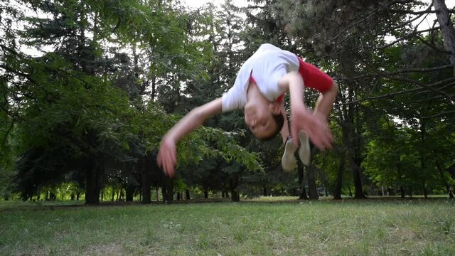 active healthy small kid in red shorts and white jersey making slow motion somersault in green park during leisure activity