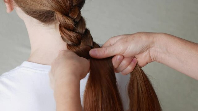 Close up view of making braid on red hair girl. Mother braids for her daughter. Process of making hairstyle for daughter. Process of braiding of red hair.