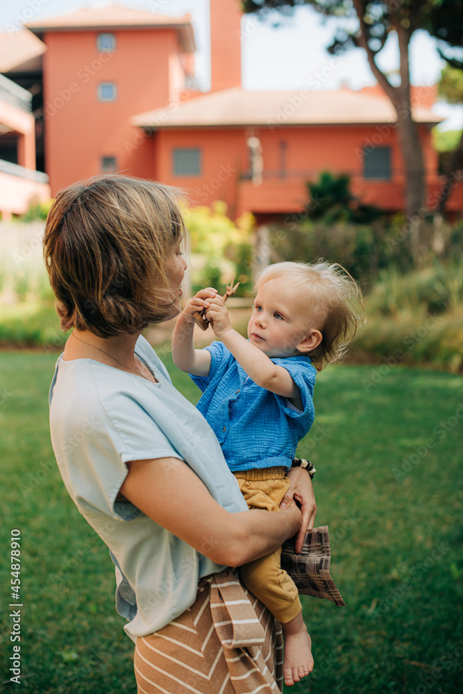 Wall mural Happy young mother walking with little daughter. Woman carrying child, toddler showing toy to her. Motherhood concept