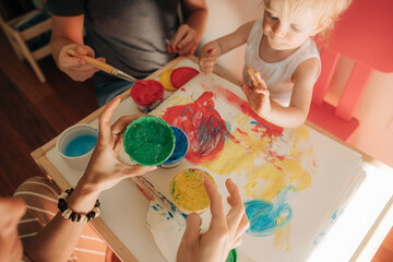 Little girl painting gouache on paper with parents. Toddler sitting with dirty hands by her mom and...