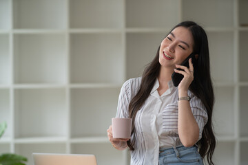 Fototapeta premium A young Asian businesswoman holding a coffee cup and talking on the phone happily at the office.