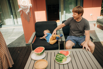 Happy father playing with little daughter outdoors. Mid adult man sitting in chair at terrace and...