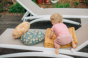 Rear view of little girl sitting on chaise lounge. Toddler resting on pillow on home terrace....