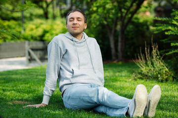 Happy young caucasian man in casual clothing sitting on grass in city park resting during a break from work, relaxed, breathes fresh air, closed eyes. 