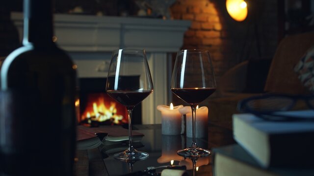 Glass of red wine, book and candle on table in dark room with fireplace. Cozy warm home in winter.