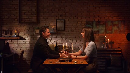 Elegant couple having romantic dinner at home, sitting at table in living room drinking red wine, talking. Stay at home concept.