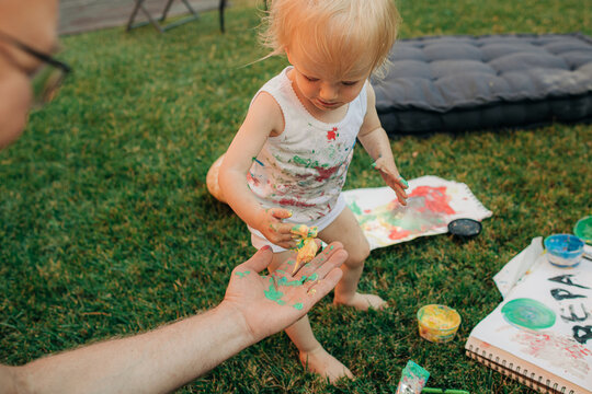 Adorable baby girl painting on mothers hand. Cute messy toddler playing with mom on front yard. Creativity concept