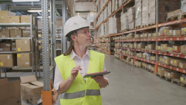 Woman manager or storekeeper in helmet walks through logistics warehouse using tablet to inventory control of goods.