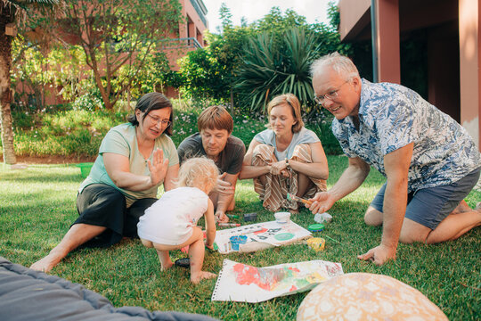 Happy multi generation family playing with child outdoors. Parents and grandparents sitting on grass and painting together with toddler. Family leisure and creativity concept