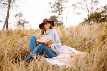 Naklejka na ściany i meble Fall picnic in nature. Young woman sitting on the blanket reading a book and smiling outdoors. People, lifestyle, relaxation and vacations concept.