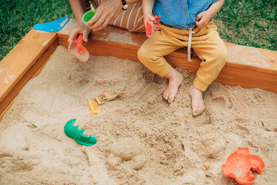 Close-up of little child sitting in sandbox with mother. Toddler kid playing in sandpit with mom outdoors. Childcare and family leisure concept