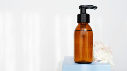  brown glass cosmetic bottle with dispenser  for soap or shampoo on blue podium with flowers on white background with sunlight. Beauty product Copy space