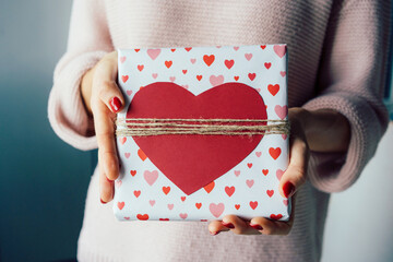 Close-up of a gift box decorated with a red heart in the hands of an unrecognizable woman.