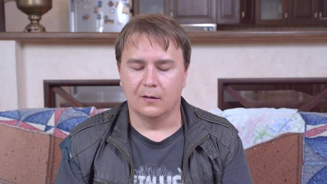 Young man in leather jacket sits on the sofa and dances slightly, shaking his head, front view. He is not happy with what is happening, so he rolls his eyes and sighs.
