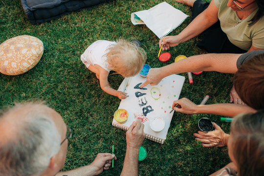Family teaching little girl how to write her name outdoors. Parents and grandparents painting with toddler on grass. Creativity and education concept