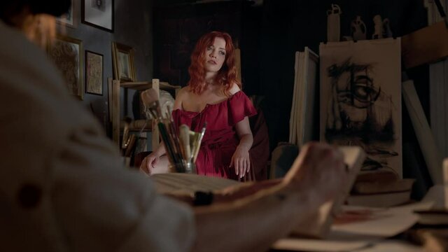 Pretty Woman With Long Red Hair Posing For Artist In Studio. Hands Of Painting Artist. Face Invisible.