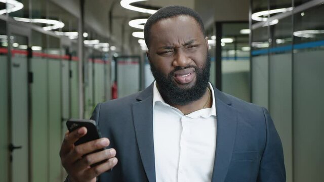 Confused African American businessman looks into a smartphone. Shocked male office worker reads an unpleasant message on a mobile phone, disgust reaction, standing in the office