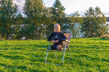 girl sunbathes on sun lounger. Background green grass trees and pond. Black jacket and pants. Blonde.