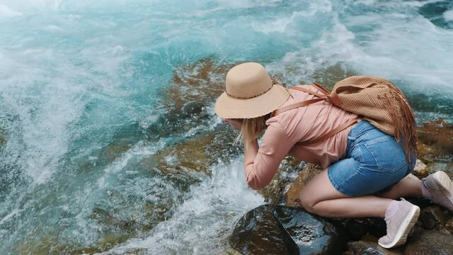 A woman traveler in a hat and with a backpack bent down to drink clean transparent water from a seething mountain river
