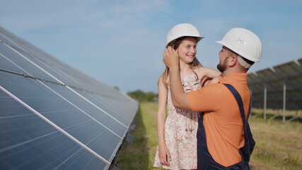A father, a solar power engineer, and his daughter are standing near solar panels. The father...