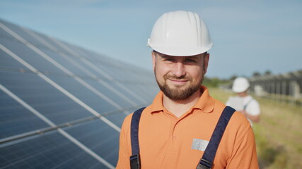 Happy caucasian adult engineer in helmet smiling at camera at solar power station outside....