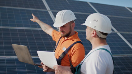 Close up engineers use laptop and project plan on the solar farm in special uniform standing look around discuss the installation of sunny batteries electricity environmental. Green energy jobs