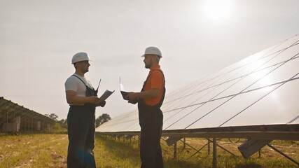 Team of industrial colleagues using tablet monitoring rows of photovoltaic solar panels at sunset. Business cooperation. Solar park. Alternative energy concept. Construction solar power plant.