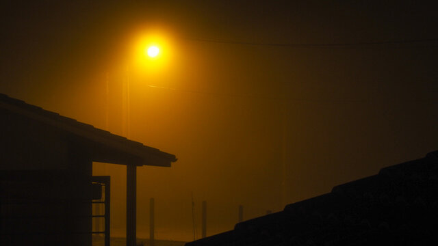Silhouettes de maisons enfouies sous un brouillard intense.   Seule la lumière intense d'un lampadaire tranche avec le reste