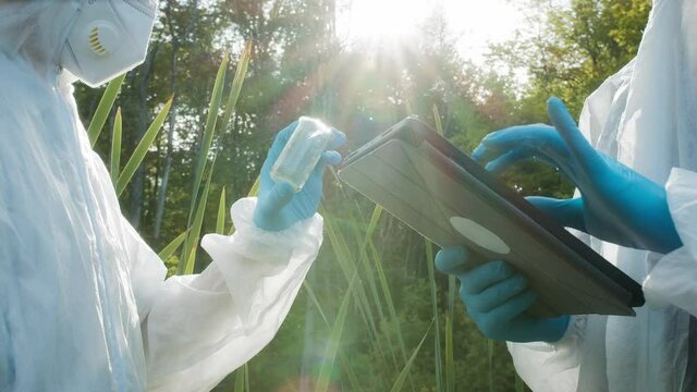 Chemists wearing ppe suits taking sample of water for toxicity testing, typing in gadget tablet. Scientific researching. Biologists in protective uniforms, field laboratory, water poisoning.