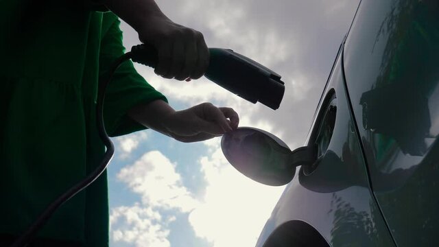 Female hands opens charging port lid an electric car and plugging in power supply cable to charge EV. Unrecognizable woman in green dress replenishes energy of vehicle on background of cloudy sky