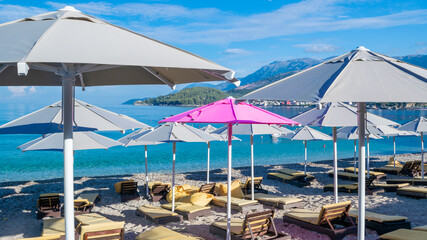 Fototapeta na wymiar One pink open beach umbrella among the rest of grays. Large beach umbrellas over sun loungers on pebble beach.