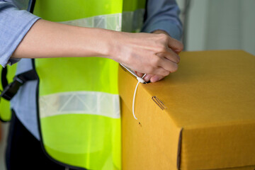 Hand of woman in a safety shirt is using a white plastic seal to lock the brown paper box to prevent the document box being illegally opened.