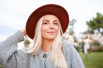Beautiful blonde smiling adult girl 20-24 year old wear casual warm clothes and hat posing in city park. Autumn season. Romantic lady outdoors. Looking at camera.