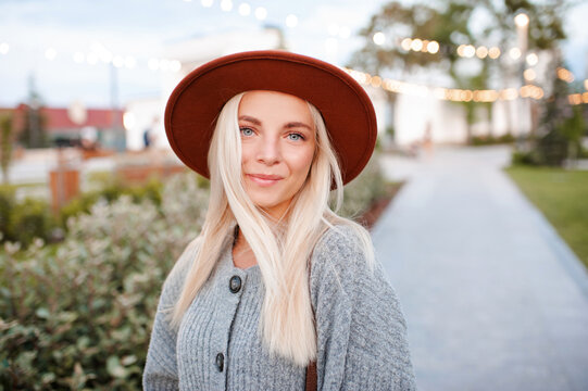 Beautiful blonde smiling adult girl 20-24 year old wear casual warm clothes and hat posing in city park. Autumn season. Romantic lady outdoors. Looking at camera.