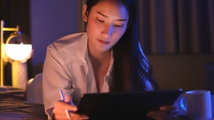 Young Asian beautiful woman enjoy using smartphone- tablet  enjoy  social online and facetime and listening music on bed with happy and smile in bedroom at night time.Depressed woman  working.