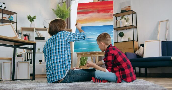Likable skilful confident bearded father-artist and his 10-aged son drawing picture together on canvas with paintbrushes at home