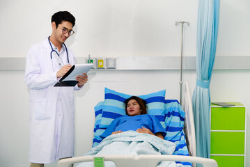 Male doctor in white gown uniform holding chart and talking to a pregnant woman laying on bed in hospital room with care and happy - 454946541