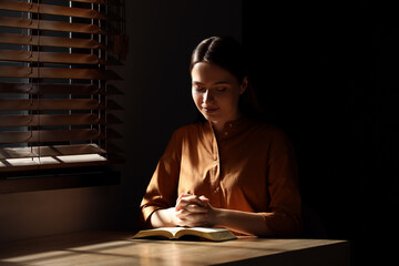 Religious young woman praying over Bible at wooden table indoors